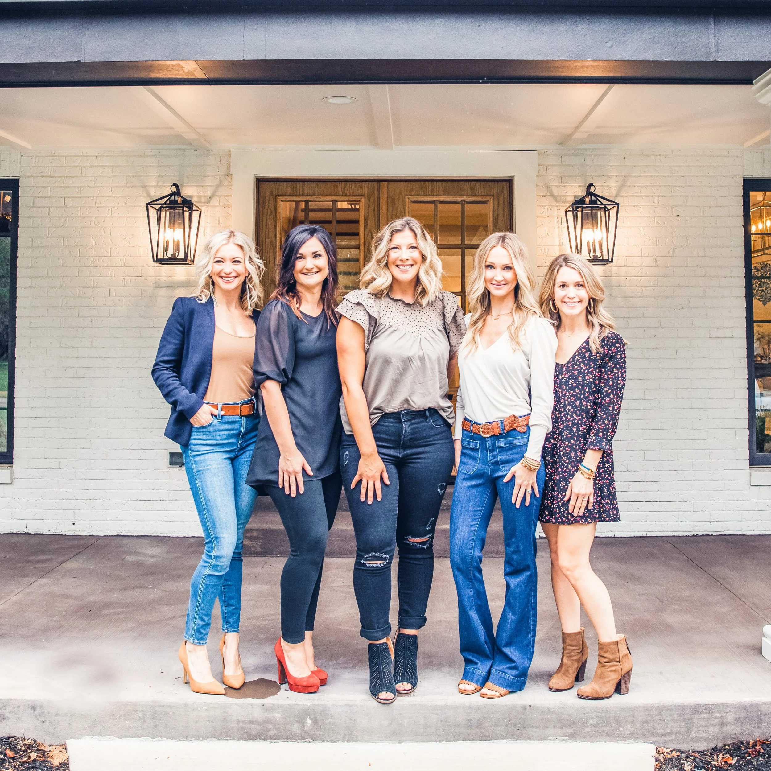 Five women working in home staging and interior design in St. Louis standing on a porch, smiling, in front of a white brick house with wooden doors and decorative lanterns.
