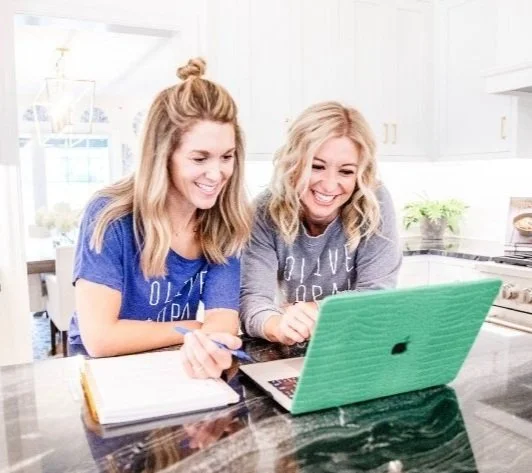 Two home staging business owners in St. Louis women and looking at a laptop in a kitchen, one holding a pen and notebook.