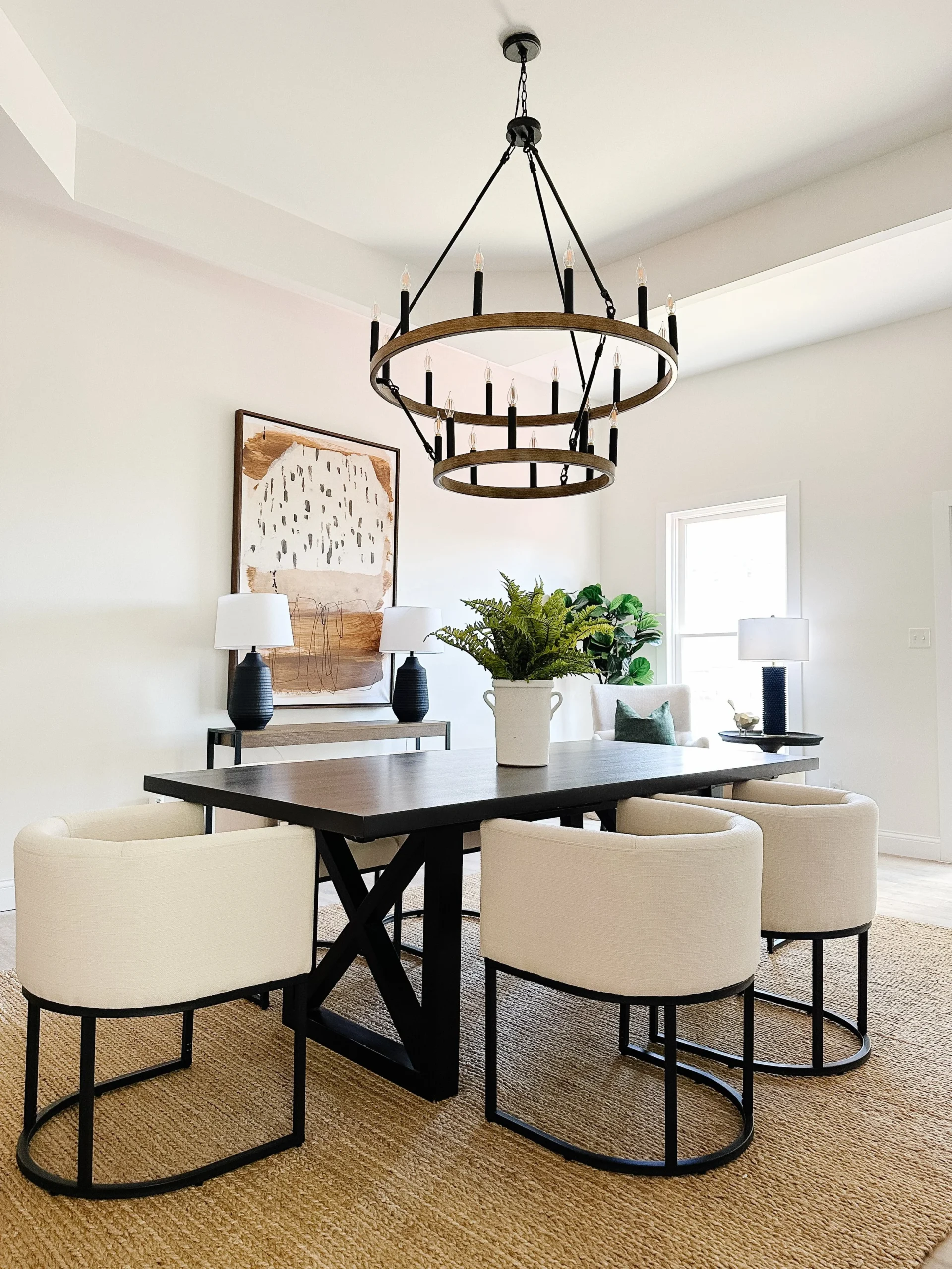 Modern dining room with a rectangular dark wooden table, surrounded by beige cushioned chairs with black frame. A large, round, double-tier chandelier hangs above. The room features light walls, a framed abstract art piece, a green plant on the table, and natural light from a window.